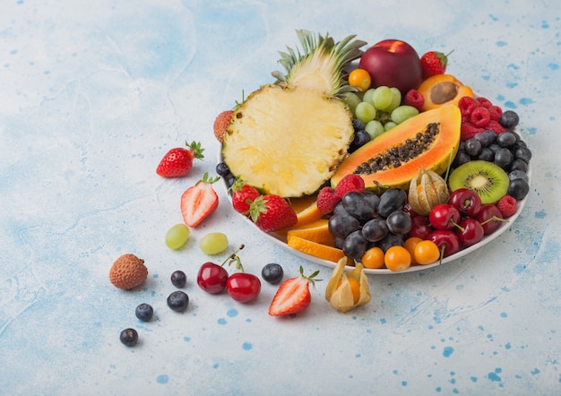 Fresh raw organic summer berries and exotic fruits in white plate on blue kitchen background. 