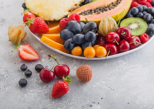 Fresh raw organic summer berries and exotic fruits in white plate on light background. Pineapple, papaya, grapes, nectarine, orange, apricot, kiwi, pear, lychees, cherry and physalis. Top view