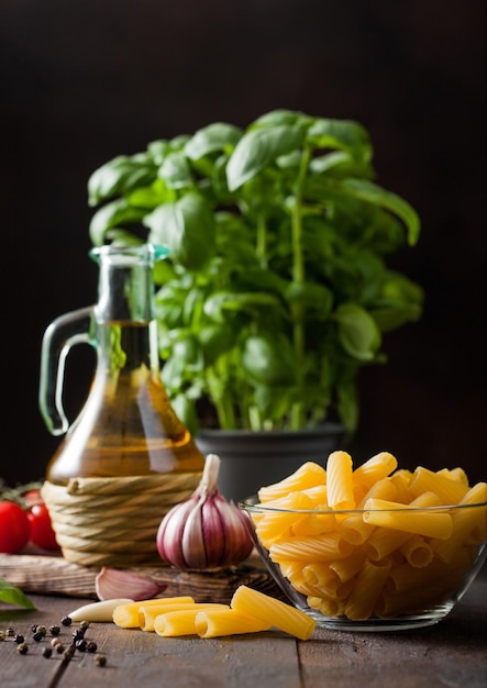 Fresh raw penne pasta in glass bowl with basil plant oil and tomatoes with garlic and pepper on wooden table background