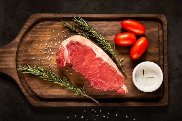 Fresh raw steak on wooden board on a dark background with salt tomatoes and rosemary