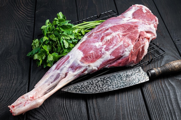 Fresh Raw whole lamb mutton leg thigh on kitchen table with herbs Black Wooden background Top view