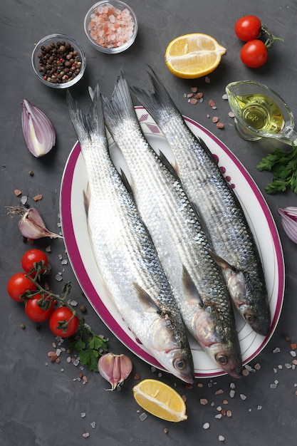 Fresh ready to cook raw mullets with ingredients and seasonings like parsley, lemon, cherry tomatoes, red onion and olive oil on dark surface