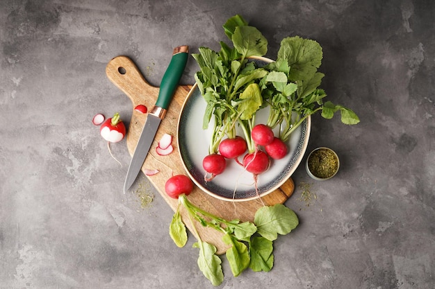 Fresh red radish with leaves on in a plate and cutting board on grey background flat lay Top view