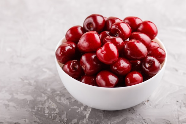 Fresh red sweet cherry in white bowl on gray concrete background. side view.