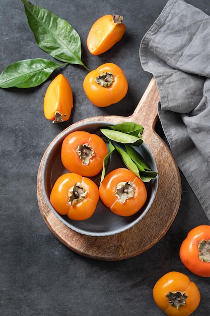 Fresh ripe persimmon fruits in bowl on a cutting board on a dark background with slices and leaves The concept of healthy nutrition and vitamins Top view