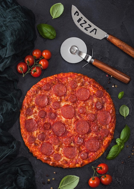 Fresh round baked Hot and Spicy Pepperoni pizza with wheel cutter and knife with tomatoes and basil on black kitchen table background