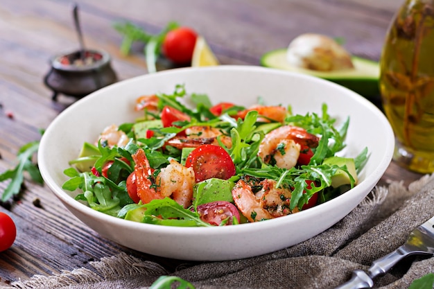 Fresh salad bowl with shrimp, tomato, avocado and arugula on wooden background close up.