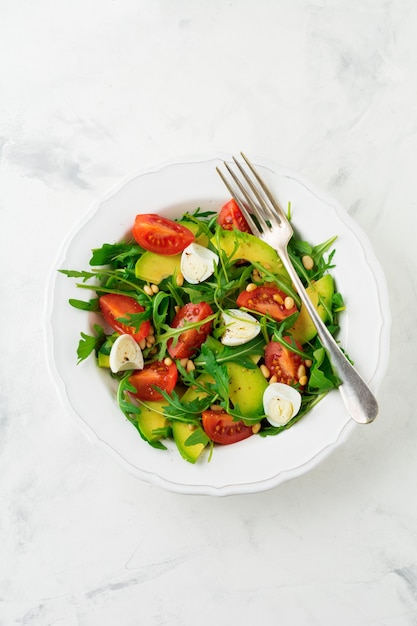 Fresh salad from arugula, tomato, avocado, olive oil, nuts and quail eggs on a light stone background