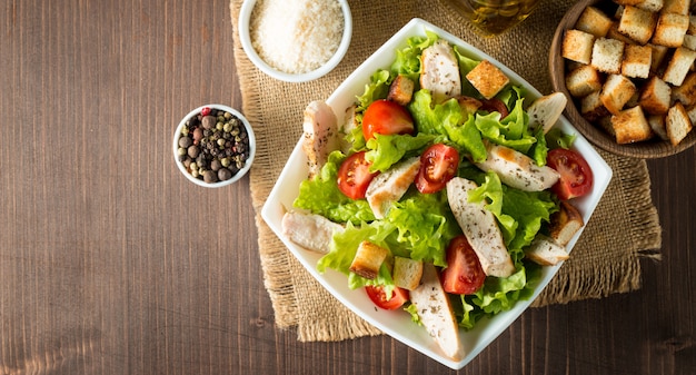 Fresh salad made of tomato, ruccola, chicken breast, eggs, arugula, crackers and spices. Caesar salad in a white, transparent bowl on wooden background