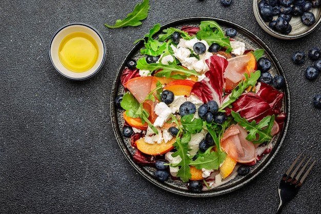 Fresh salad with arugula radicchio sweet peaches smoked pork ham feta cheese and blueberries Black stone table background top view