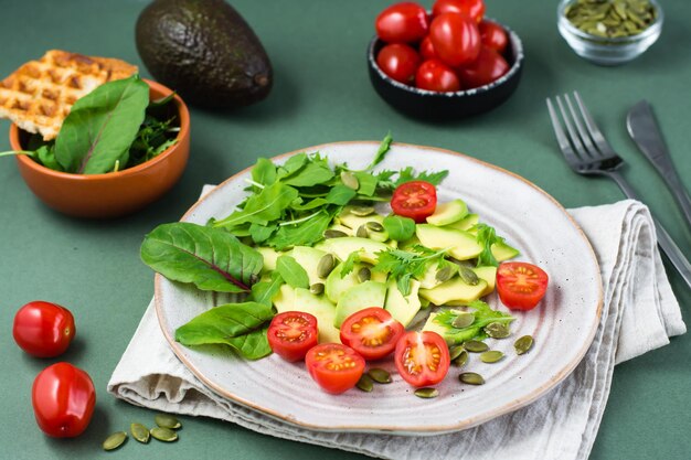 Fresh salad with avocado, cherry tomatoes, arugula, mizuna, chard and pumpkin seeds on a plate on a green background. Vegetarian vitamin food.