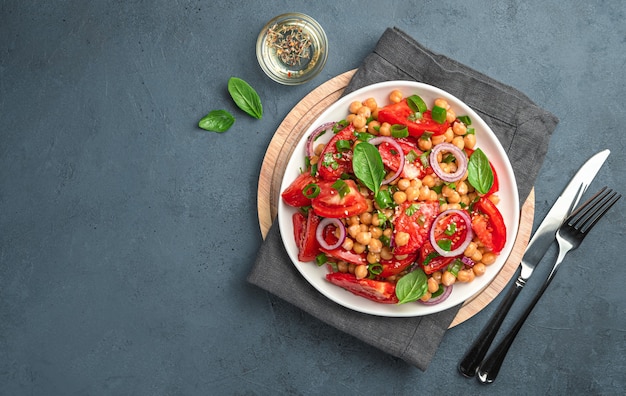 Fresh salad with tomatoes, chickpeas, onions, basil and olive oil on a dark gray-blue background. Top view, space for copying.