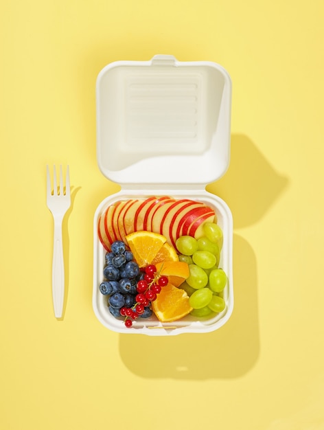 Fresh sliced fruit and berries in white take away box isolated on yellow background, top view