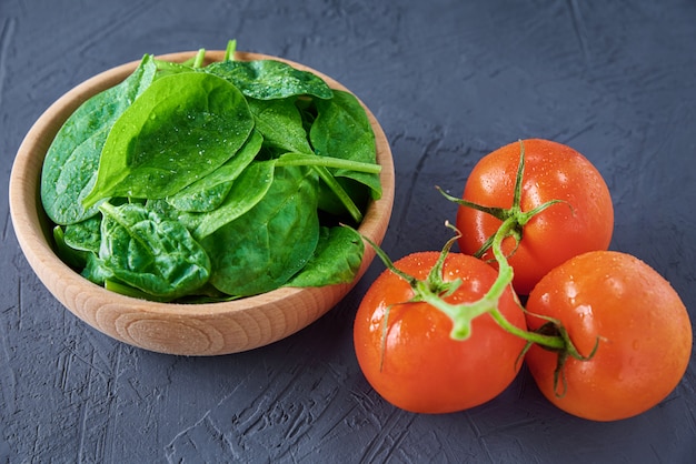 Fresh spinach leaves in wooden bowl and tomato on a dark background