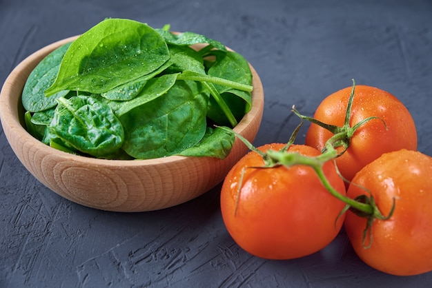 Fresh spinach leaves in wooden bowl and tomato on a dark background.