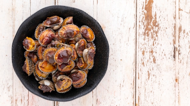 Fresh steamed cockles, boiled cockles in black ceramic plate on white wood texture background with copy space for text, top table view, flat lay, blood cockle