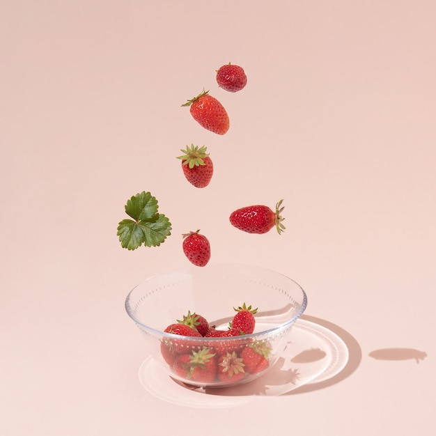 Fresh summer fruits, strawberries, and green leaves fall into a glass bowl, isolated on a pink background. Creative food layout. Square