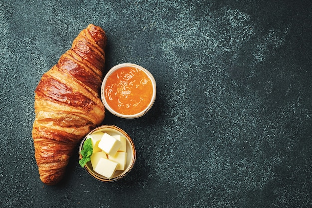 Fresh sweet croissant with butter and orange jam for breakfast. Continental breakfast on a black concrete table. Top view with copy space. Flat lay.