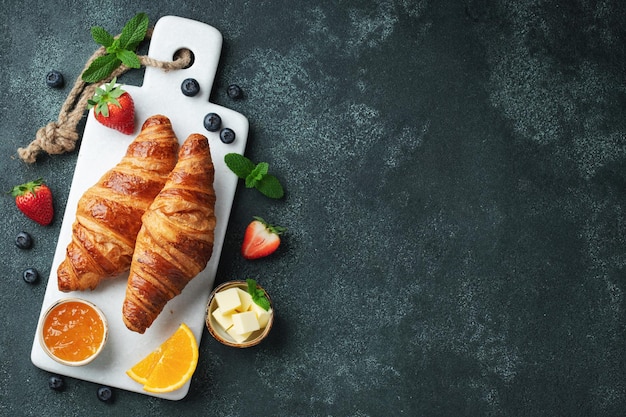 Fresh sweet croissants with butter and orange jam for breakfast Continental breakfast on a dark concrete background Top view with copy space Flat lay