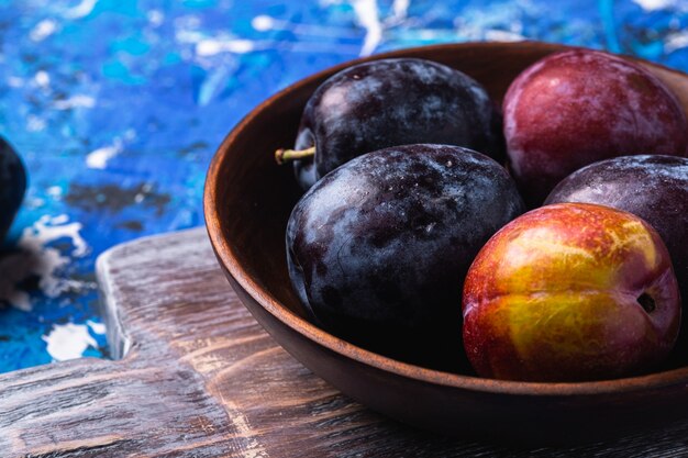 Photo fresh sweet plum fruits in brown wooden bowl on old cutting board, blue abstract table, angle view macro