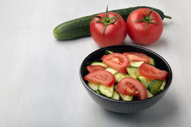 Fresh tomato and cucumber salad in dark bowl on white wooden table