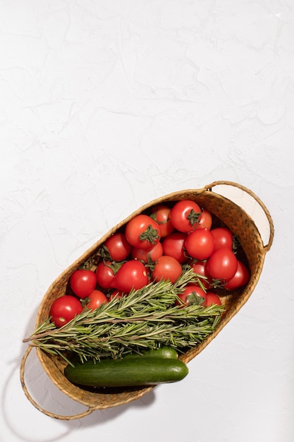 Fresh tomatoes cucumbers and a few sprigs of rosemary in a wicker basket on a white surface