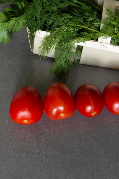 Fresh tomatoes and dill greens with parsley in a basket of vines lie on a gray table