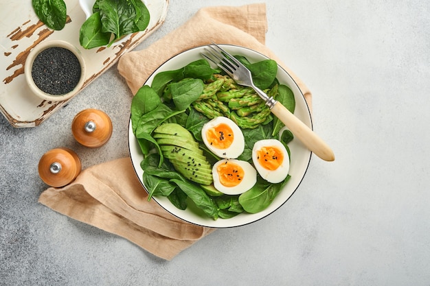 Fresh vegetable salad with avocado, asparagus, crumpled eggs with black sesame seeds and spinach on plate on light slate, stone or concrete background. Balanced lunch in bowl. Top view. Mock up.