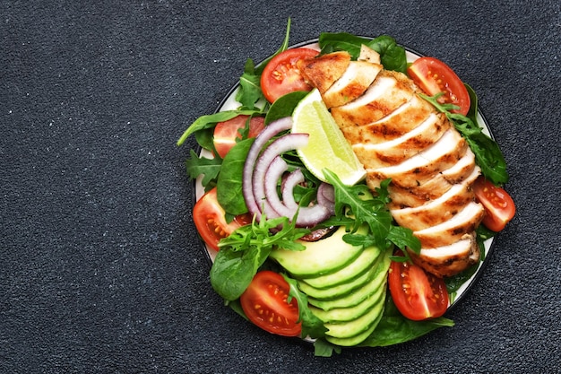 Fresh vegetable salad with grilled chicken fillet spinach tomatoes avocado and red onion with olive oil on black background Healthy detox ketogenic diet food Top view