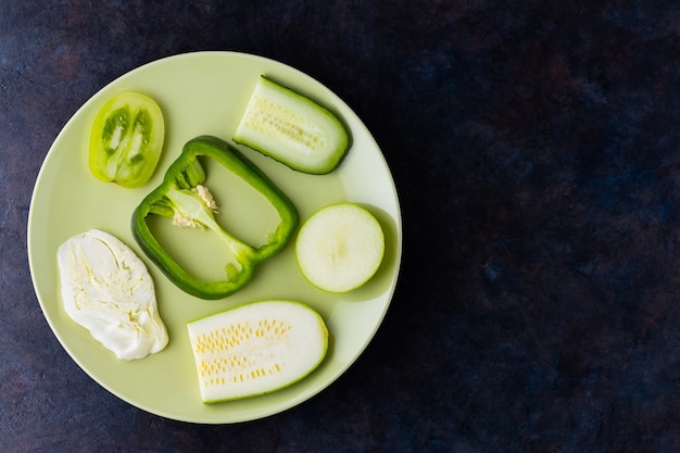 Fresh vegetables and fruits on a green plate. Various vegetables on a dark wall. Bell peppers, tomatoes, cucumber, cabbage, zucchini and apple for vegan. Top view. Copy space