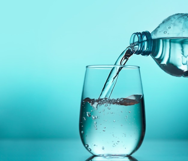 Fresh water pouring from the plastic bottle into a glass on the blue background. Drink water each day is important for optimal health