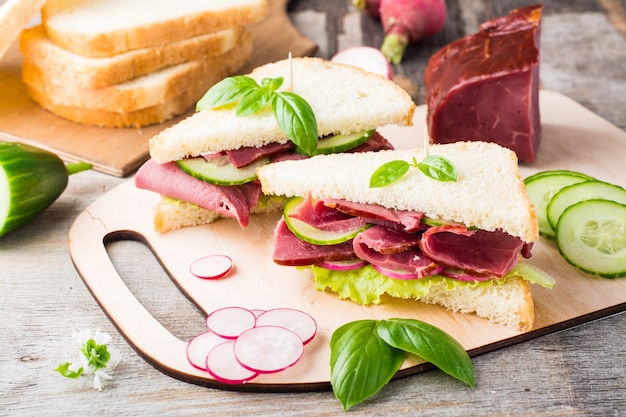 Fresh white bread sandwiches with pastrami, cucumber, radish and basil on a cutting board. American snack. Rustic style