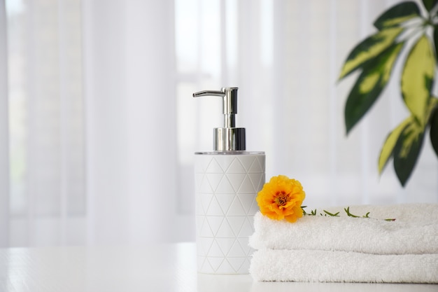 Fresh white towels folded on white table, orange flower and liquid container with green leaves of house plant and tulle window on background.