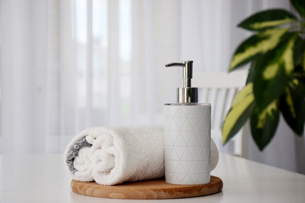 Fresh white towels rolled on wooden board and liquid container with green leaves of house plant and tulle window on background. Copy space.