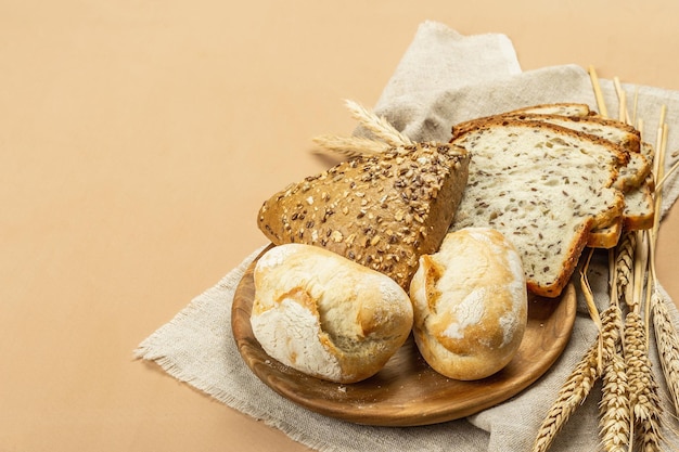 Freshly baked bread with wheat ears, buns, and toasts. Assorted bakery on light beige background. Vintage linen napkin, wooden plate, copy space