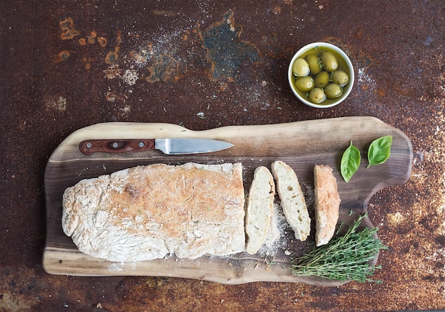 Freshly baked homemade ciabatta bread with olives, basil and thyme on walnut wood board over grunge rusty metal , top view