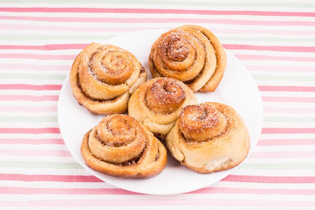 Freshly baked homemade snail buns with sugar and cinnamon on white plate and striped tablecloth. Balanced nutrition, proteins and carbohydrates, cereals