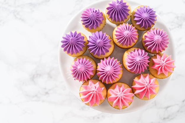 Freshly baked mini vanilla cupcakes with ombre pink buttercream frosting on a white cake stand