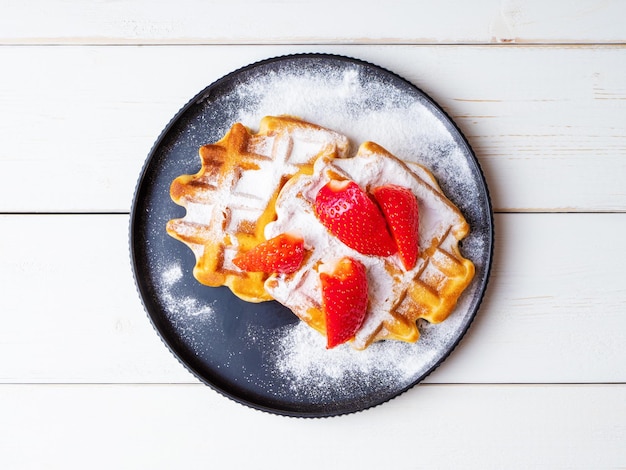 Freshly baked Viennese waffles sprinkled with powdered sugar with strawberries lying on a black ceramic plate on a wooden background Top view