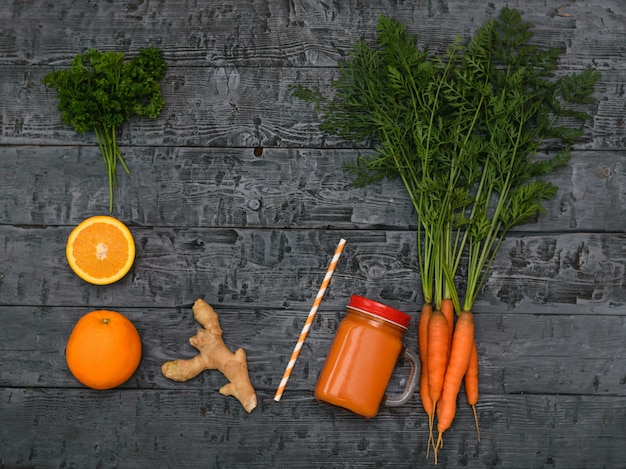 Freshly made carrot smoothie, bunch of carrots, orange and ginger root on a rustic dark table.