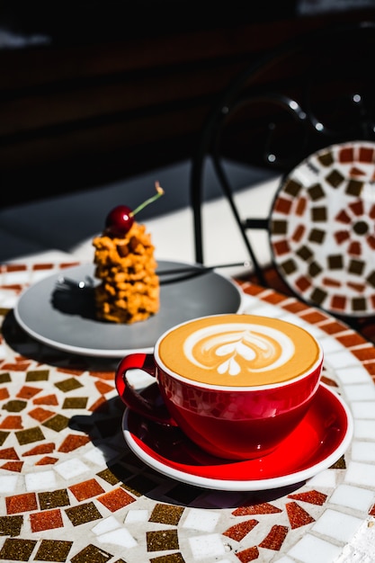 A freshly made red cup of cappuccino coffee and a dessert on a mosaic stone table of a cafe terrace on a sunny day