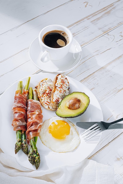 Fried asparagus wrapped in jamon fried eggs bruschetta with soft cheese and avocado on a white plate and a cup of freshly brewed coffee