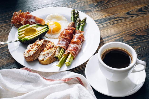 Fried asparagus wrapped in jamon fried eggs bruschetta with soft cheese and avocado on a white plate and a cup of freshly brewed coffee