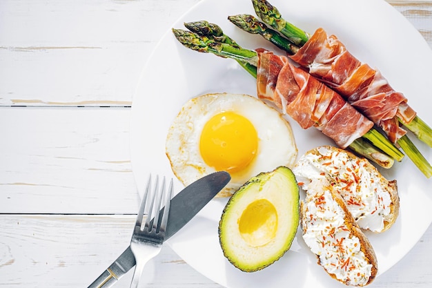 Fried asparagus wrapped in jamon fried eggs bruschetta with soft cheese and avocado on a white plate