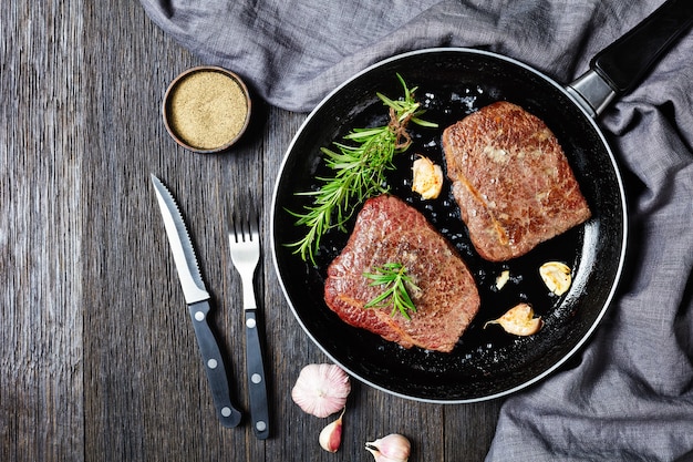 Fried beef steaks with rosemary bouquet, garlic and salt on a skillet on a wooden table with fork and knife, horizontal view from above, flat lay, free space