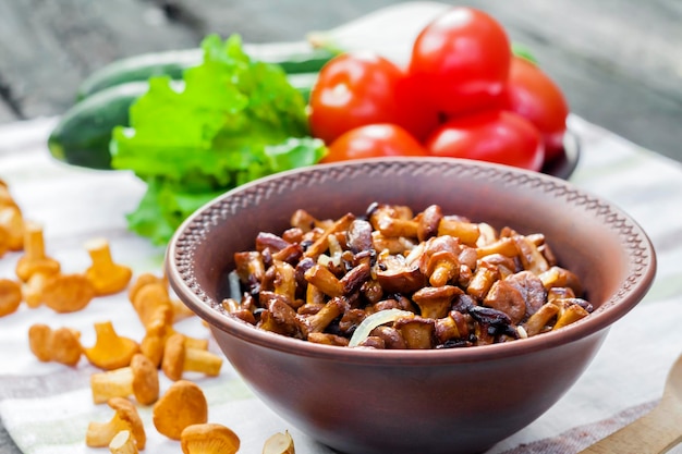 Fried chanterelles with onion in rustic bowl and plate with fresh vegetables for salad on background