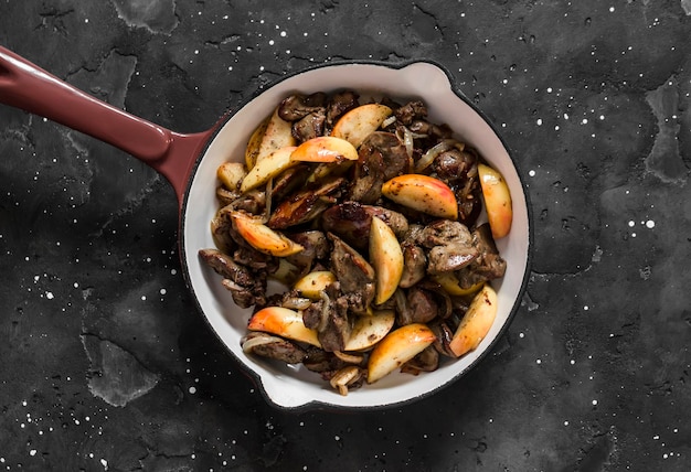 Fried chicken liver with onions and apples in a ceramic frying pan on a dark background top view