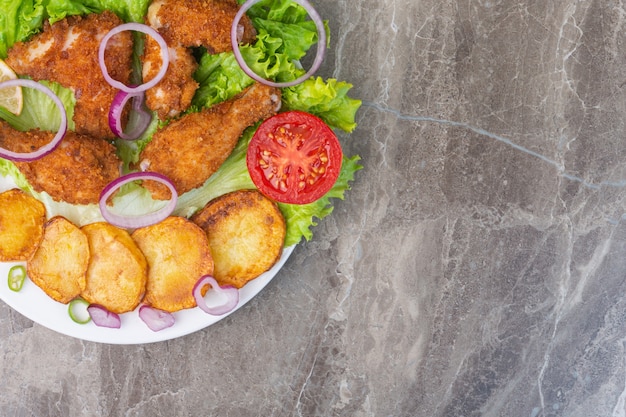 Fried chicken meat, potatoes and vegetables on a plate, on the marble background.