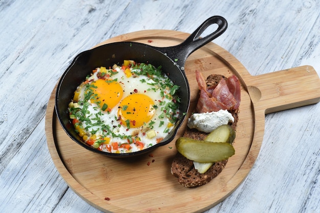 Fried eggs in a pan with vegetable tartare, bacon, pickled cucumber and cream cheese. top view