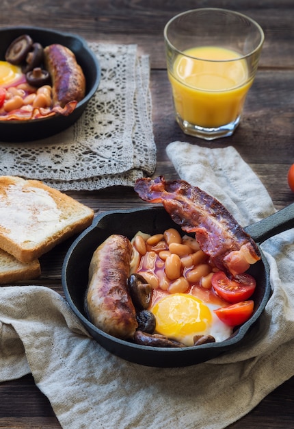 Fried eggs, sausages, bacon, beans and mushrooms in iron skillet, toasts, orange juice, butter and jam on rustic wooden background. Full english breakfast. Selective focus.